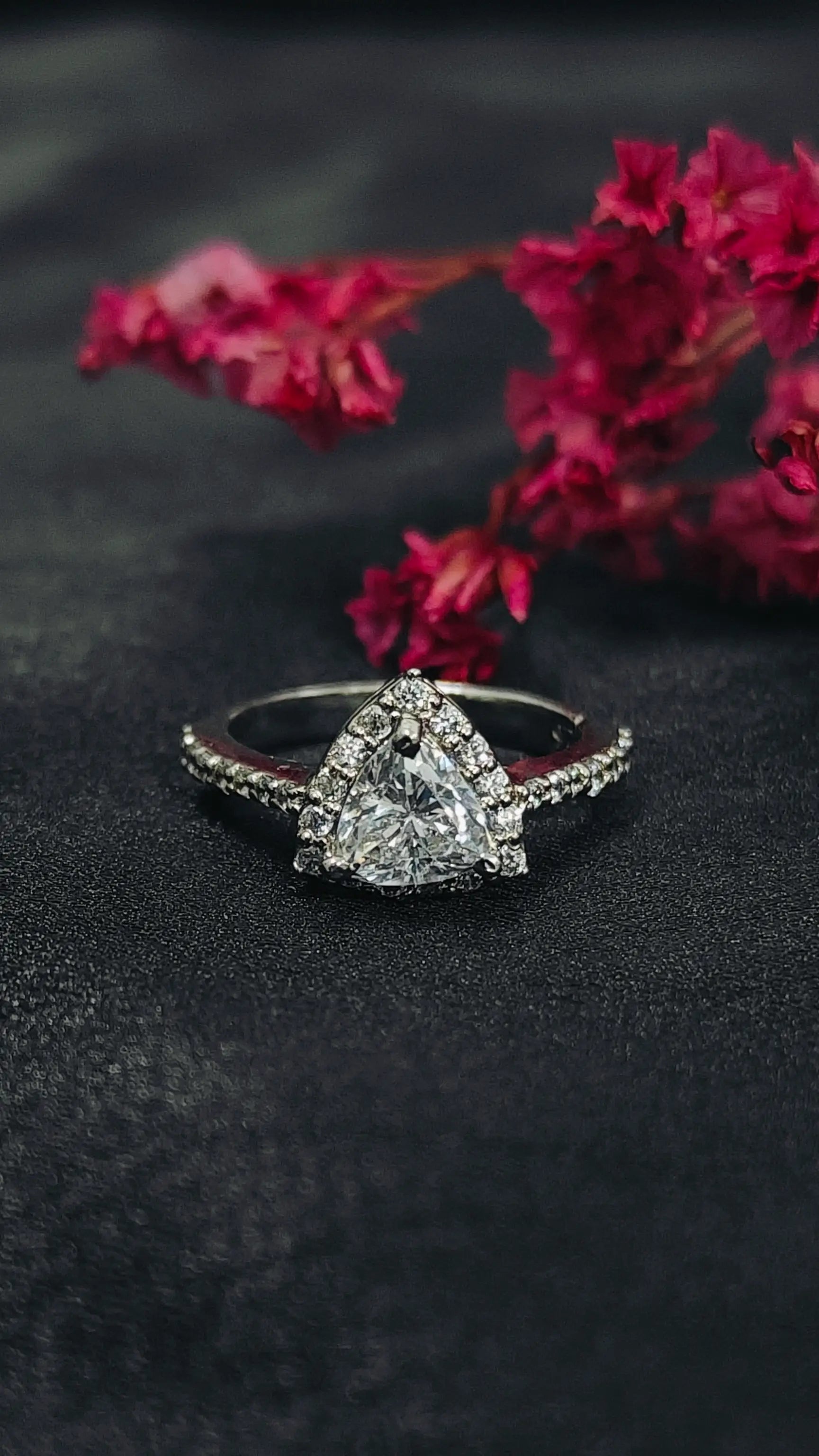 Diamond ring with a triangle-shaped center stone on a dark surface with pink flowers.